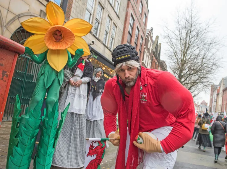 “Person dressed in a red Welsh rugby outfit posing next to a large daffodil decoration during St David’s Day celebrations on a city street