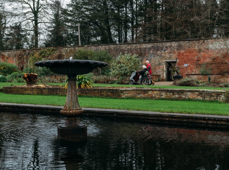 A woman in a motorised scooter and a man walking in an ornamental garden.