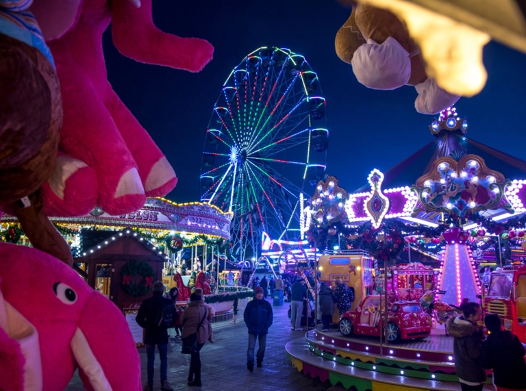 night time shot of winter wonderland fairground with toys in the foreground, people walking between amusements and a big wheel lit up in the background.