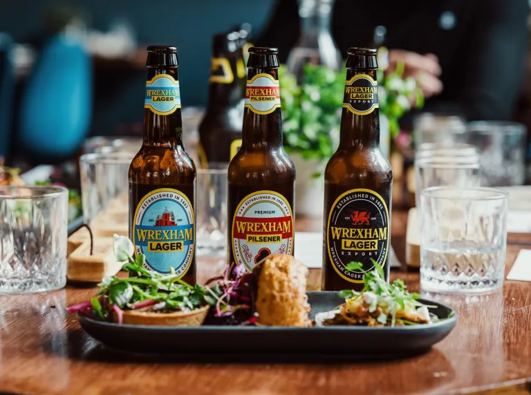 Close-up of a wooden table with craft beer bottles and a plate of gourmet appetizers, including greens and artisanal bites, set in a stylish dining setting