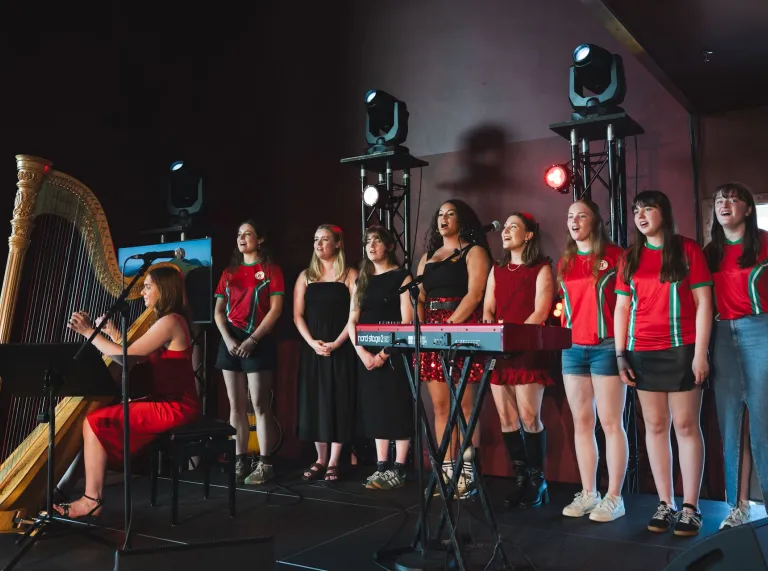 A harpist performing on stage alongside a group of young women standing in a row, dressed in coordinated red and black outfits, with stage lights and a keyboard in the background
