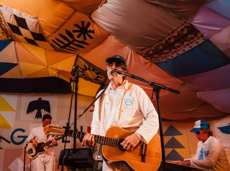 Musician performing on stage with a guitar, wearing a white outfit and cap, under a colourful patchwork canopy, with band members playing instruments in the background.