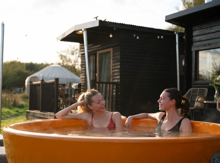 Two people sitting in a round, bright orange outdoor hot tub at a rustic retreat. Behind them is a small dark wooden cabin with a pitched roof and a white tent structure in the background. The setting includes grassy terrain and natural greenery under soft daylight, creating a relaxed outdoor spa atmosphere.