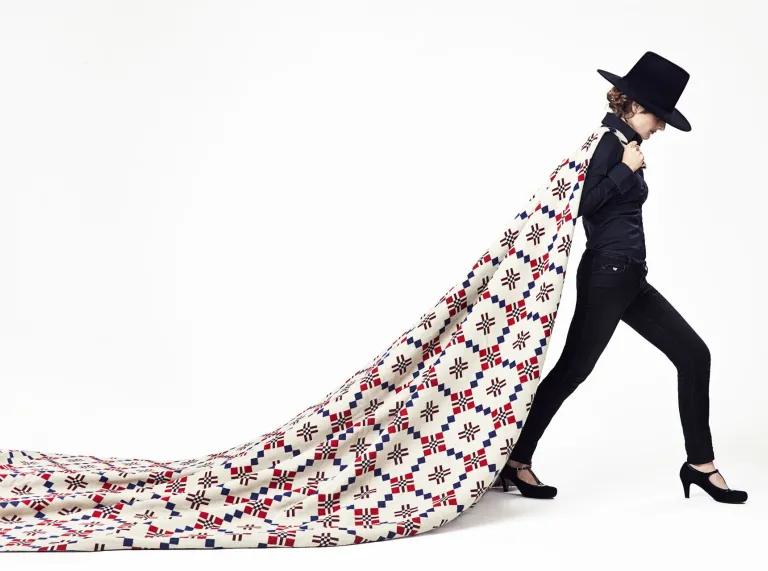 Person dressed in an all-black outfit with a wide-brimmed hat and high-heeled shoes, walking forward while dramatically pulling a large patterned quilt behind them