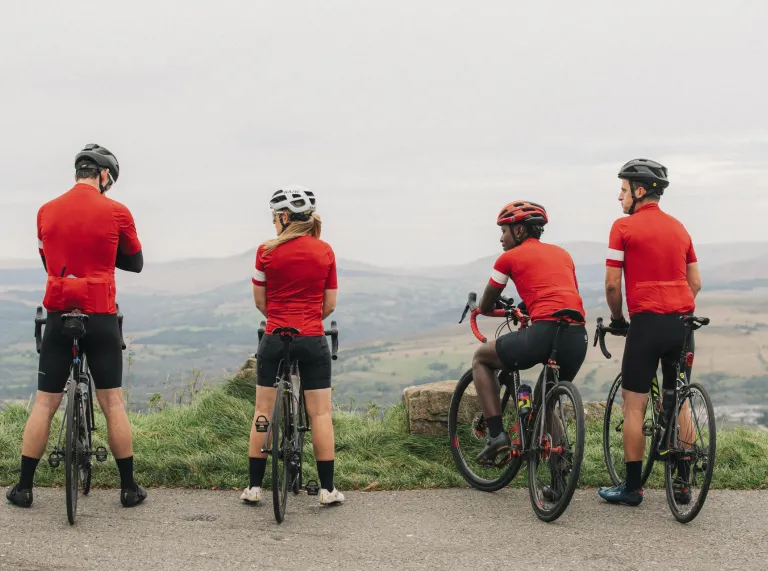 A group of four cyclists wearing red jerseys pause with their bikes at a viewpoint overlooking rolling hills and countryside.