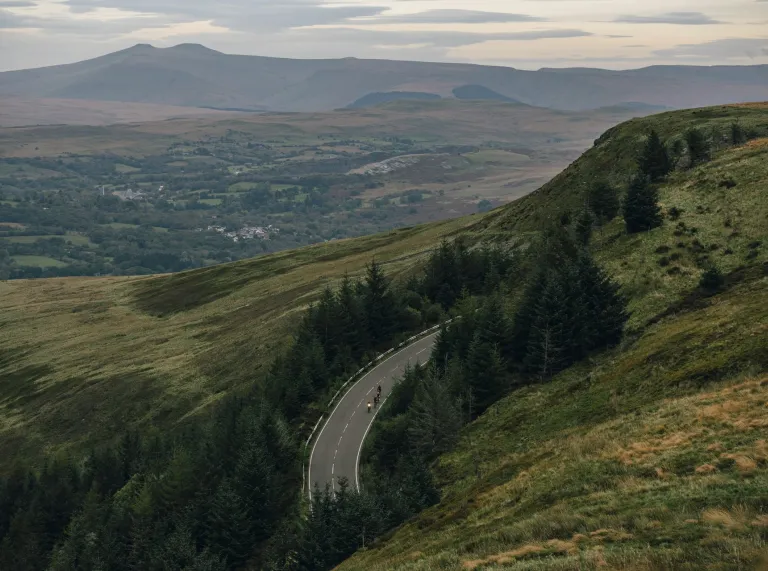 A winding mountain road cuts through green hills and dense trees, with a small group of cyclists riding along it, set against a wide landscape of valleys and distant peaks