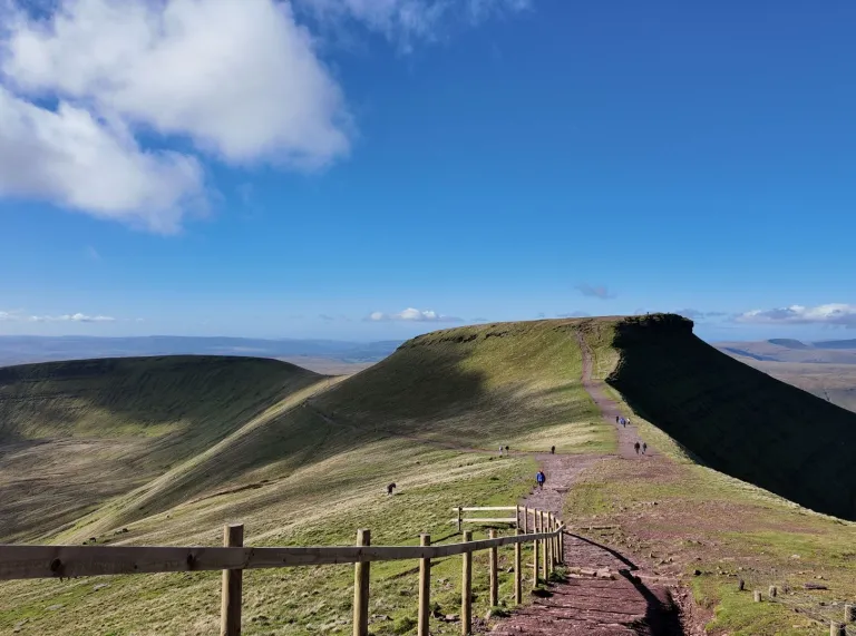 A wide mountain landscape with a clear blue sky, showing a path leading along a ridge towards a flat-topped peak, with walkers scattered along the trail and green hills stretching into the distance.