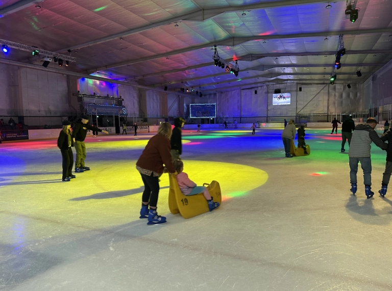 People skating in an indoor ice rink with colourful lights cast across the ice. Some adults are pushing children seated on yellow skating aids, while others skate around the rink.