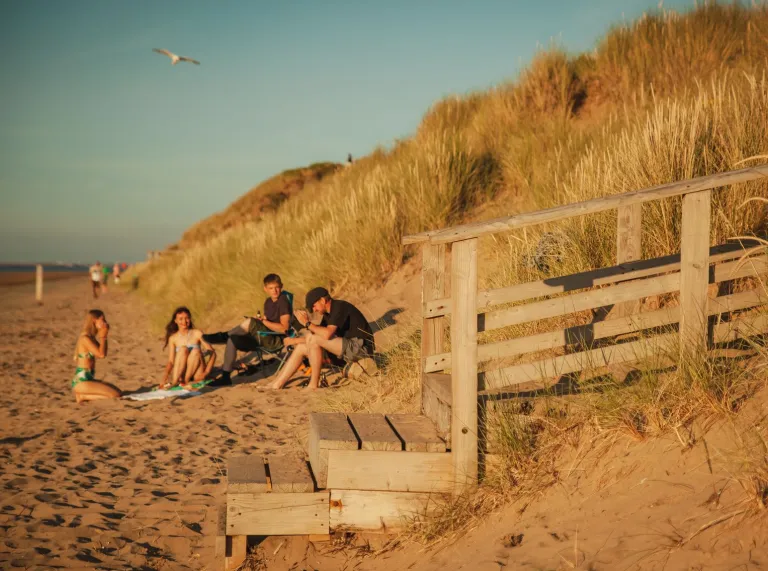 A group of people sat by sand dunes on a sandy beach.