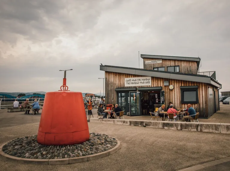 A rustic wooden beachfront café with outdoor seating, a bright red buoy sculpture in the foreground, and a cloudy sky overhead.