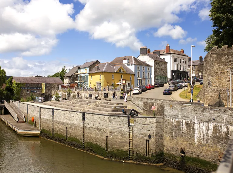 View of a riverside walkway in a historic Welsh town, with stone walls, colourful buildings on a hill, and calm water in the foreground under a partly cloudy sky.