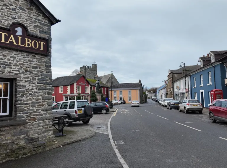 Street view of a Welsh town with stone buildings, colourful painted shopfronts, and parked cars lining a gently curving road under an overcast sky.