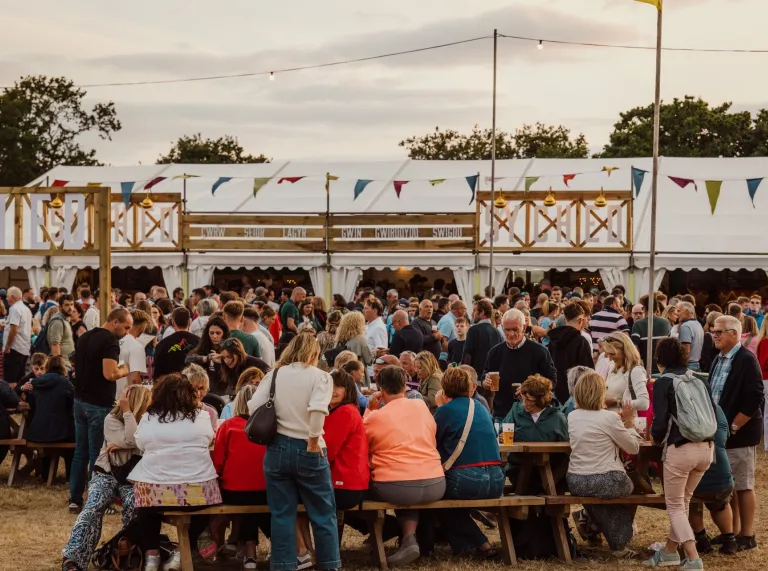 Crowds of people at an outdoor festival sat on benches outside a large marquee. 