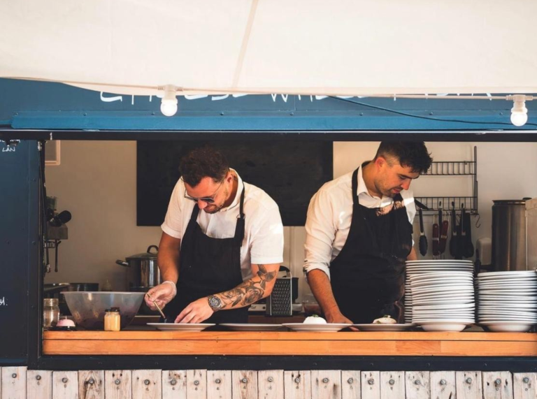 two chefs in a small open kitchen preparing food behind a wooden counter.