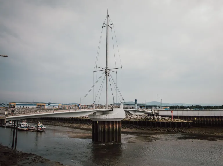 A pedestrian and cycle bridge with a tall central mast spans across a tidal river on an overcast day. Boats sit on the muddy riverbed below due to low tide, and distant hills are visible under the cloudy sky.