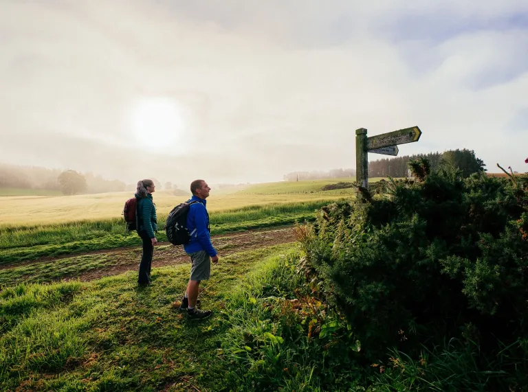 Two hikers on a countryside path, looking at a national trail waymarker wooden post.rpost.