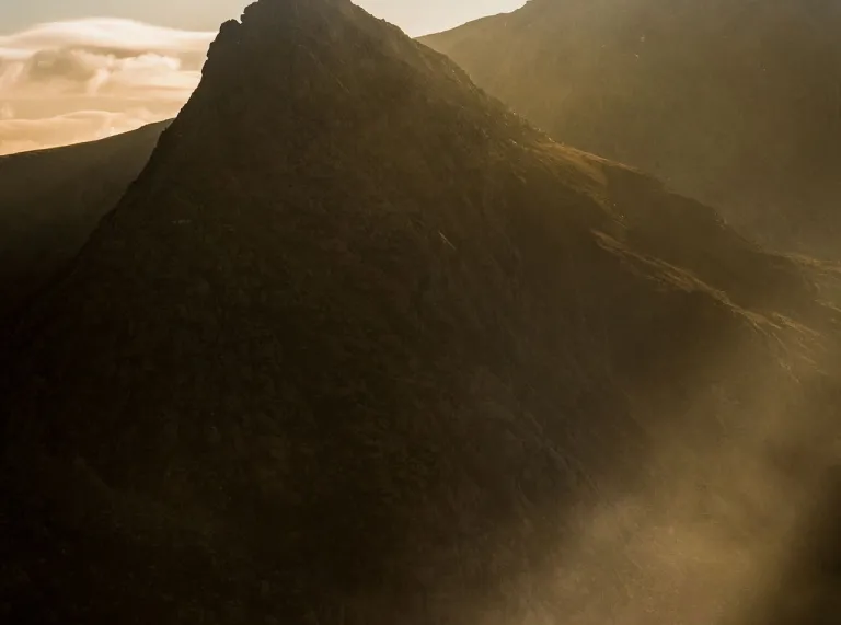 A silhouetted figure stands on a rocky ridge with a small dog, facing a steep, dark mountain peak lit by angled beams of golden light.