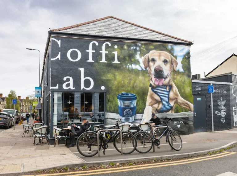 Exterior view of Coffi Lab café with outdoor seating and bicycles parked outside, featuring a large wall mural of a dog beside a takeaway coffee cup.