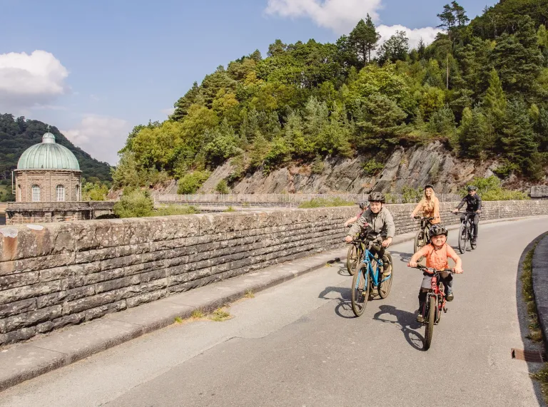 Family cycling across a stone dam with a domed water tower and wooded hills in the background.