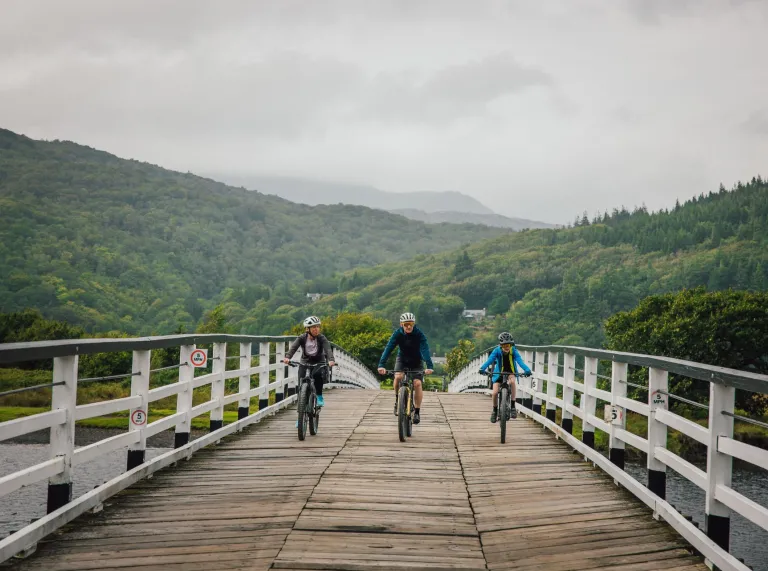 Drei Radfahrer fahren über eine breite Holzbrücke, die über einen Fluss führt, im Hintergrund sind grüne Hügel und bewaldete Hänge zu sehen.