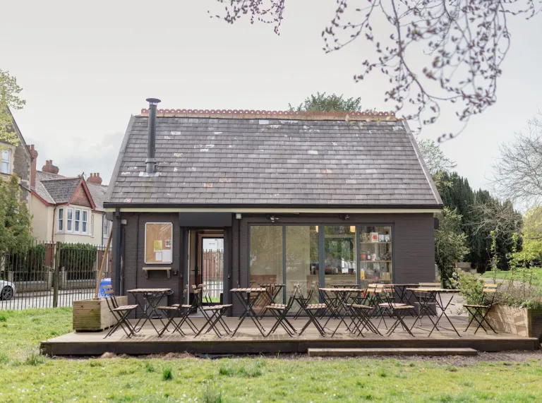 Modern small wooden cabin with a pitched roof, large glass doors and windows, and a small deck, set in a grassy garden with shrubs and neighbouring houses visible in the background.