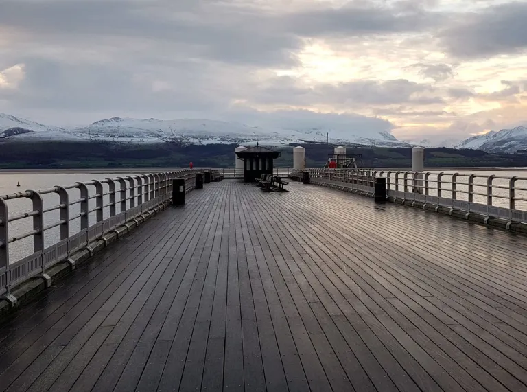 A long wooden pier reaching out over the water towards snow covered mountains in the distance.