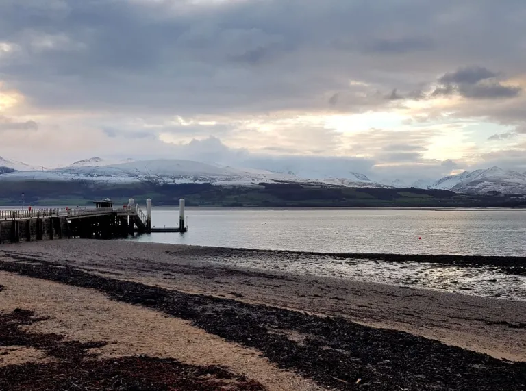 A wooden pier reaching out over sand and water with snow covered mountains in the distance.