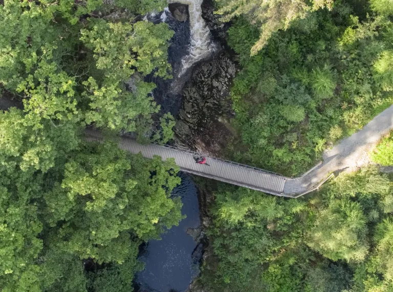 Aerial view of a footbridge crossing a river gorge surrounded by dense woodland, with a waterfall visible upstream.