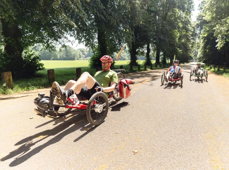Three people using sit down cycles on a wide path in a green city centre park.