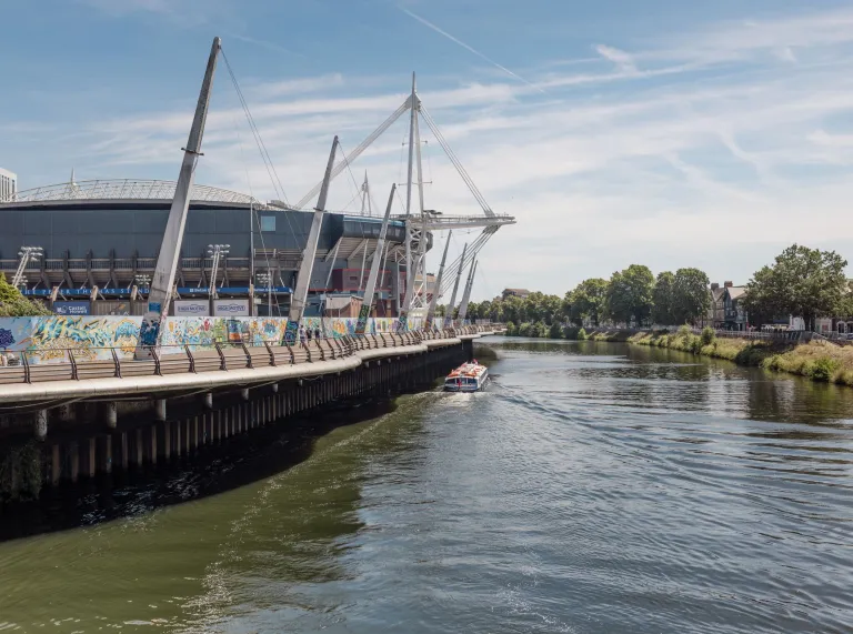 Boat travelling along the River Taff beside a graffiti-covered walkway, with the Principality Stadium and its distinctive support masts in Cardiff