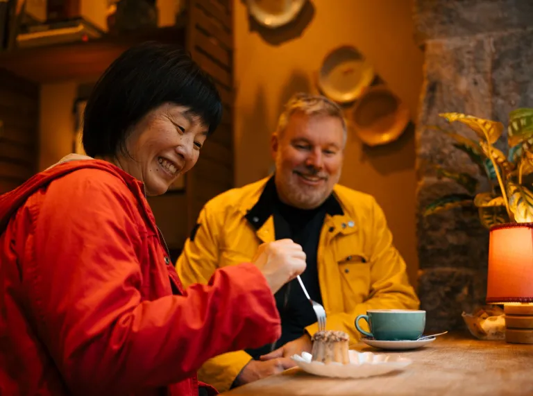 Two customers enjoying coffee and cake at a wooden table in a cosy café setting.
