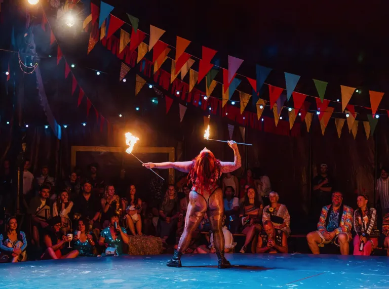 Fire performer twirls flaming batons on a circular stage inside a festival tent while spectators watch beneath colourful bunting.