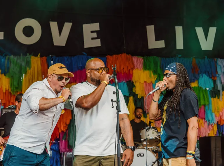 Three vocalists perform together on the Big Love Festival stage in front of a brightly coloured backdrop and live band.