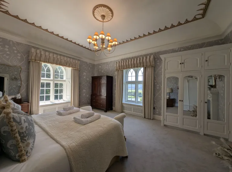 Elegant bedroom with a double bed, arched windows, fitted wardrobes, patterned wallpaper and a chandelier, with views of the surrounding countryside.