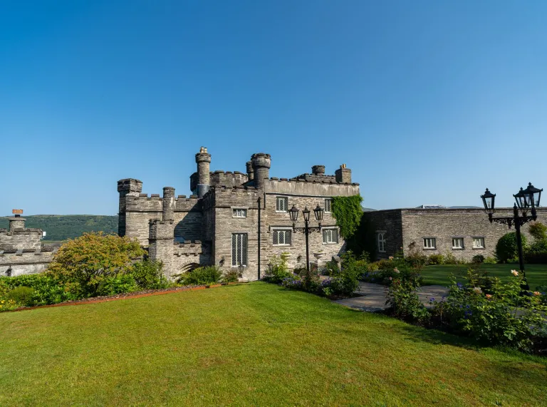 Stone-built castle surrounded by landscaped gardens and lawns, with flower borders, ornamental lamp posts and distant hills beneath a clear blue sky.