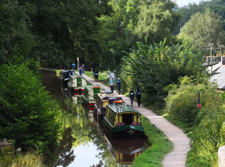 Several walkers following a countryside canal tow path, with narrowboats moored up alongside.