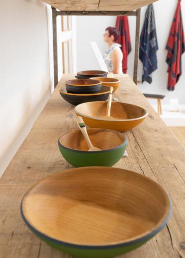 Wooden bowls and spoons on display on a shelf with fabrics hanging on a wall.