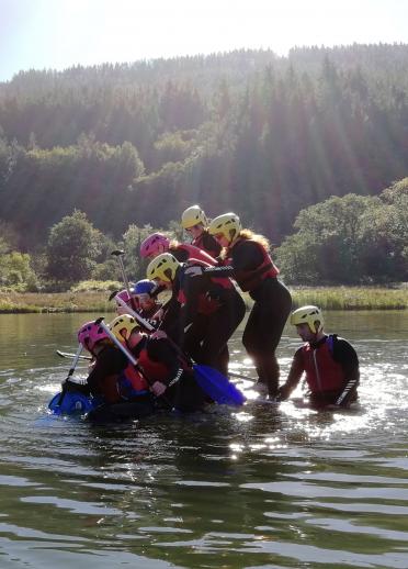 Adult group in shallow water with red life jackets and yellow safety hats, climbing onto a blue raft during a team building event 