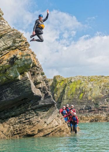 A man jumping off a cliff, coasteering into the sea with a group looking on.