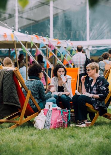 A group of ladies relaxing in deckchairs at the Hay Festival with show tents and bunting in the background.