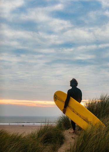 surfer holding surf board with tall grass and sea in background.