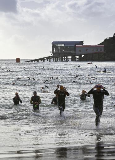 Competitiors finishing swimming stage and running onto North Beach with old and new lifeboat stations in background
