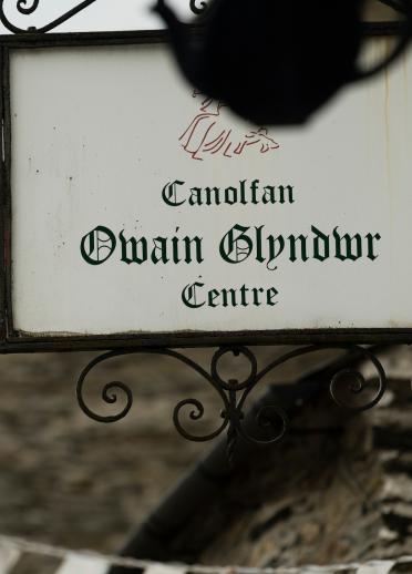 Schild des Owain Glyndŵr Centre mit der wehenden Owain Glyndŵr Fahne dahinter.