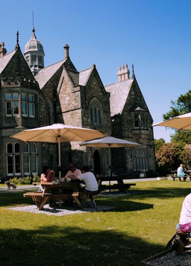 people sat at patio tables with parasols with a manor type building in the background