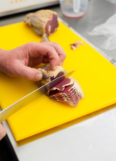 A person slicing cured lamb on a yellow chopping board in a kitchen