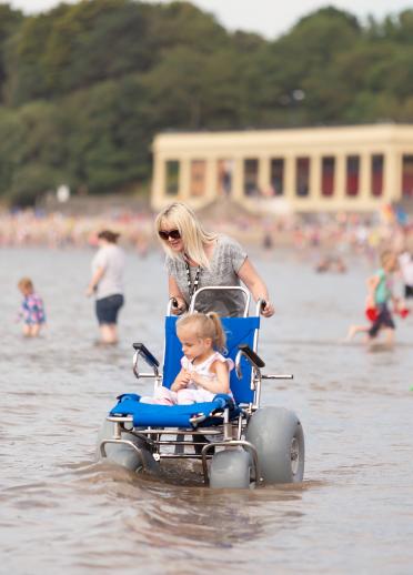 girl in sea using all-terrain beach wheelchair with mother behind.