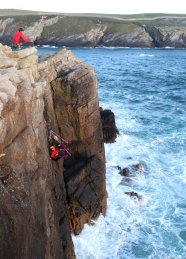 A cliff face above the sea with two people sitting on a portaledge suspended from the cliff edge with safety instructor on top of the cliff. 