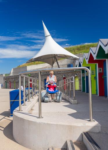 People using a beach wheelchair by brightly coloured beach huts.