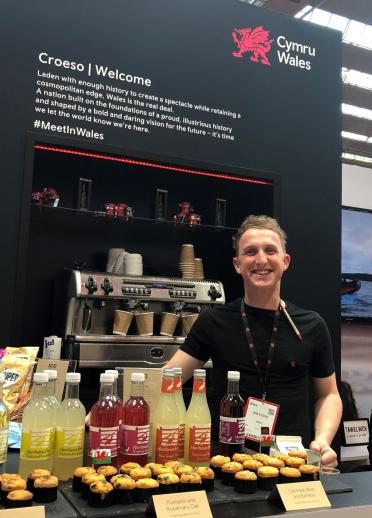 Wales male staff member with a black tshirt on standing in front of the Wales exhibition with a range of Wales food and drink.