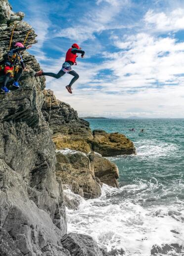 Two adults in life jackets and helmets jumping into the sea off the rocks coasteering